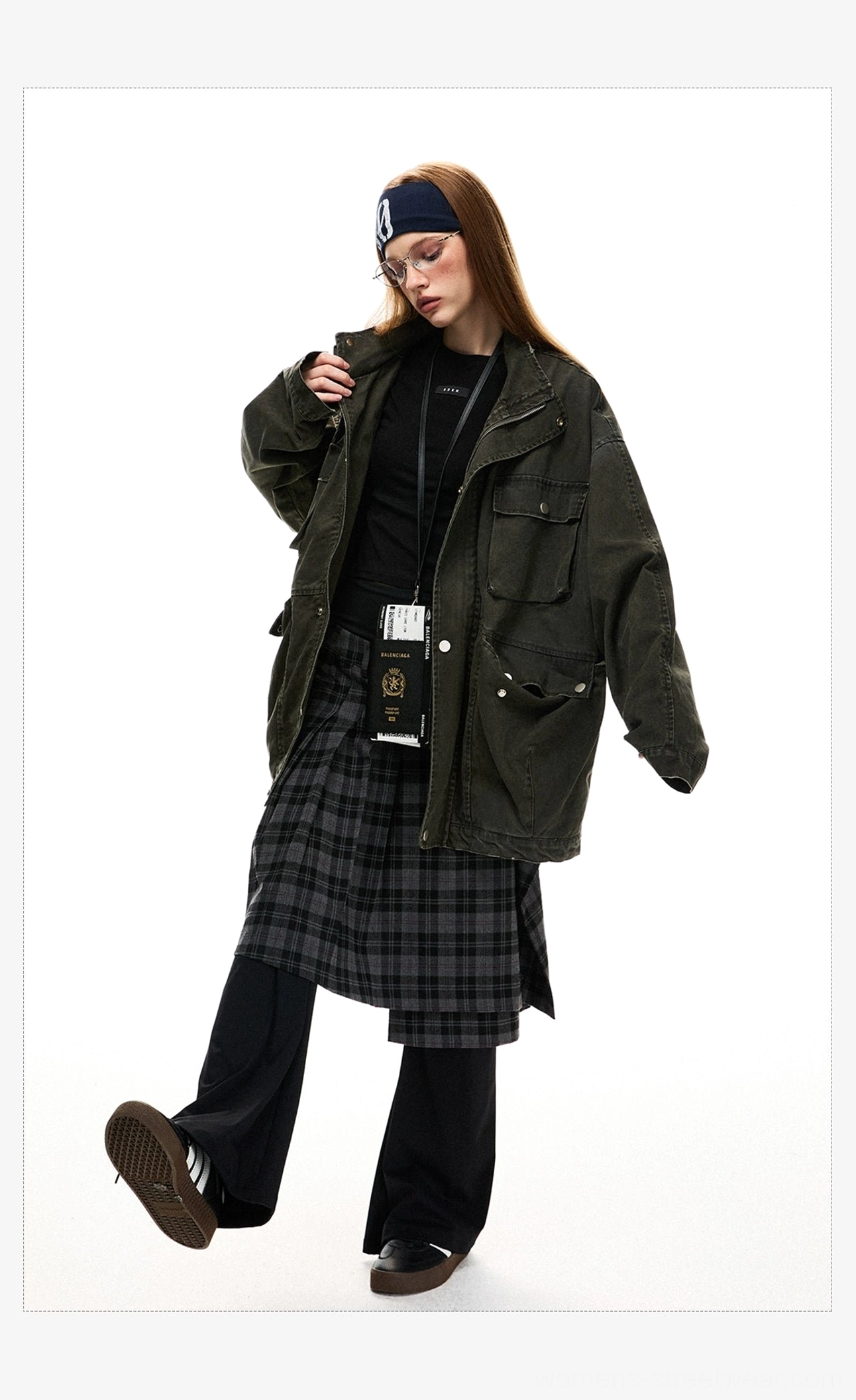 Three-quarter view of a woman styling an oversized olive green utility jacket with a plaid skirt, trousers, and crossbody streetwear accessory