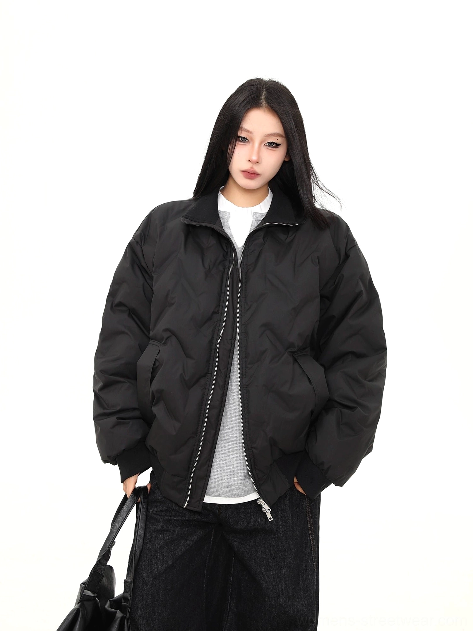 Female model wearing an oversized black chevron quilted puffer bomber jacket with a stand collar, ribbed trim, side pockets, and a black leather tote bag