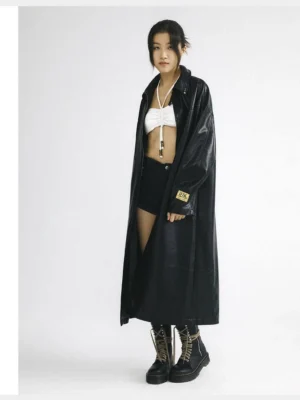 Female model wearing an oversized black faux leather hooded coat in a full-body side view with a smooth finish, relaxed sleeves, and dramatic streetwear silhouette