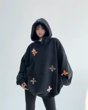 Front three-quarter view of an oversized black floral appliqué hoodie with colorful four-petal flowers, leopard-print accents, dropped shoulders, and a kangaroo pocket