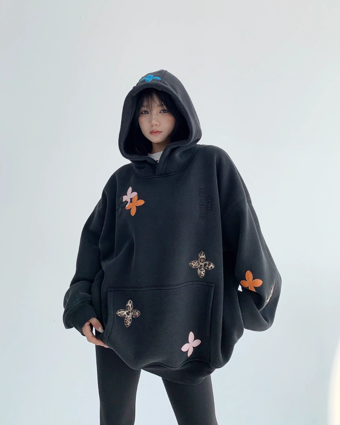 Front three-quarter view of an oversized black floral appliqué hoodie with colorful four-petal flowers, leopard-print accents, dropped shoulders, and a kangaroo pocket