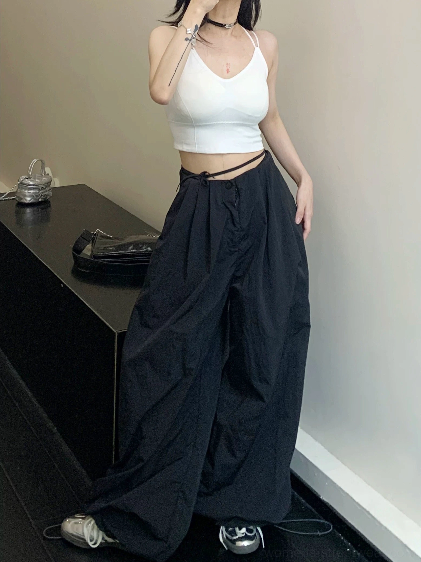 Black oversized parachute wide-leg streetwear pants styled with a white cropped camisole and retro sneakers in a minimalist studio
