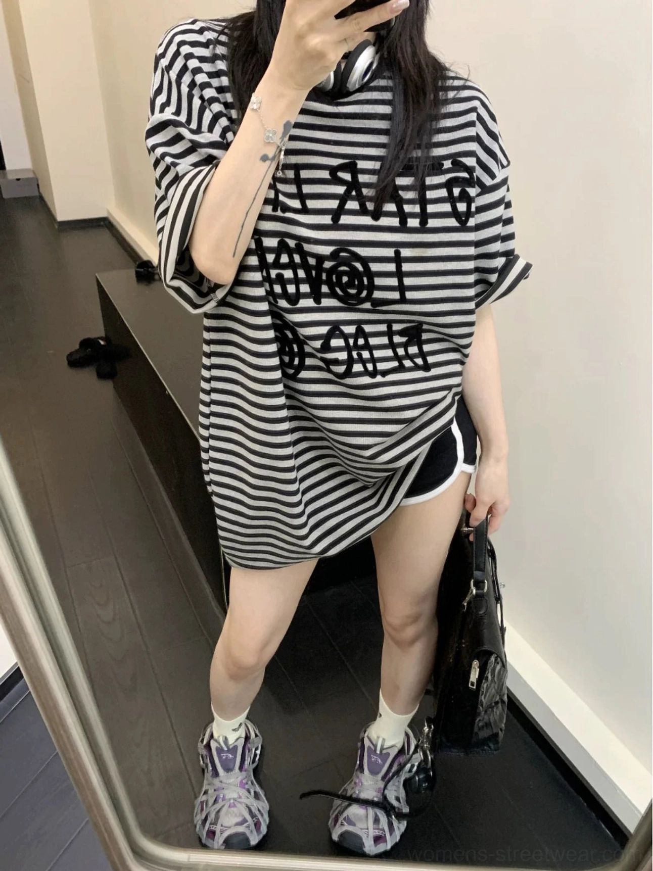 Full-body back view of a woman wearing an oversized black striped T-shirt with biker shorts, sporty sneakers, and a leather tote bag