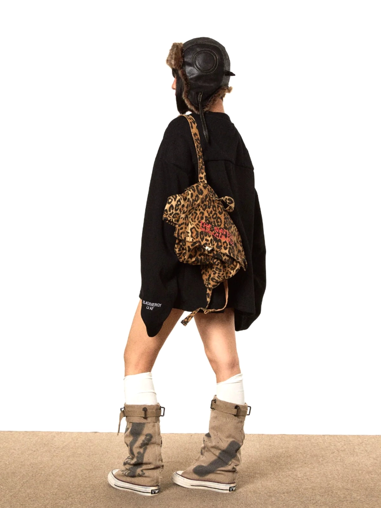Back view of a hip-covering oversized black V-neck sweater with long loose sleeves and subtle cuff prints styled with knee-high socks and work boots