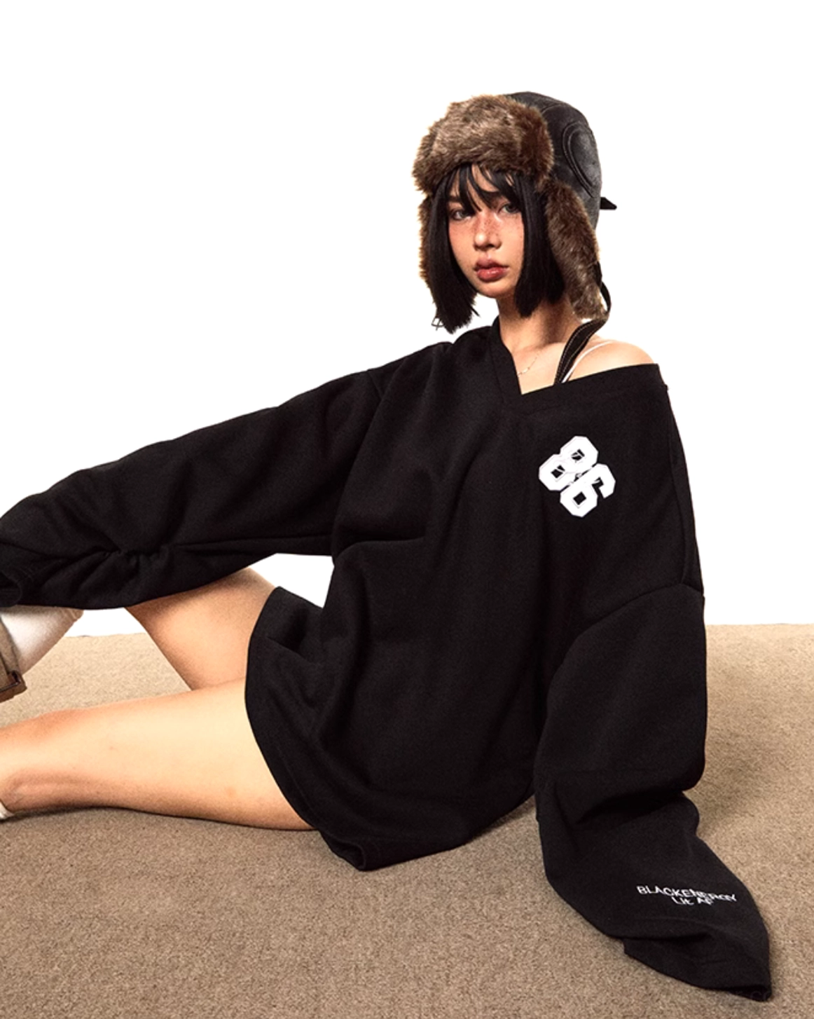Side seated view of an oversized black V-neck sweater worn off one shoulder with extra-long loose sleeves and 86 chest embroidery