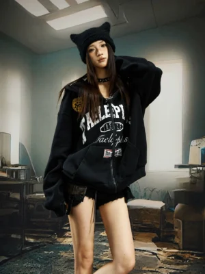 Oversized black varsity patch zip-up streetwear hoodie worn by female model in industrial warehouse setting