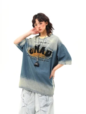 Front three-quarter view of an oversized blue distressed denim wash graphic tee with frayed chest lettering, dropped shoulders, light blue cargo pants, and pendant necklace