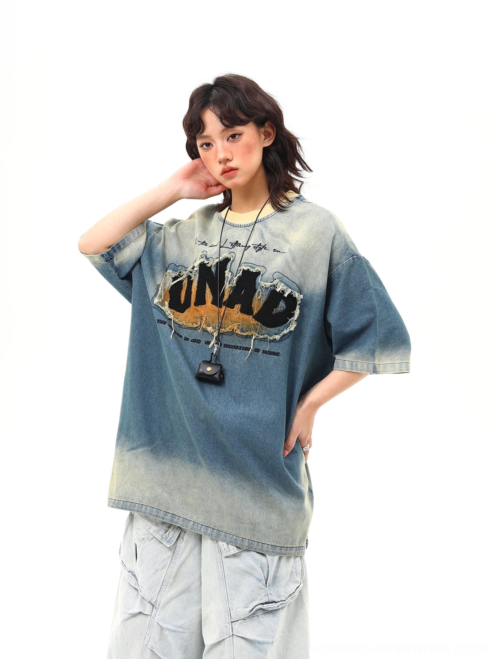 Front three-quarter view of an oversized blue distressed denim wash graphic tee with frayed chest lettering, dropped shoulders, light blue cargo pants, and pendant necklace