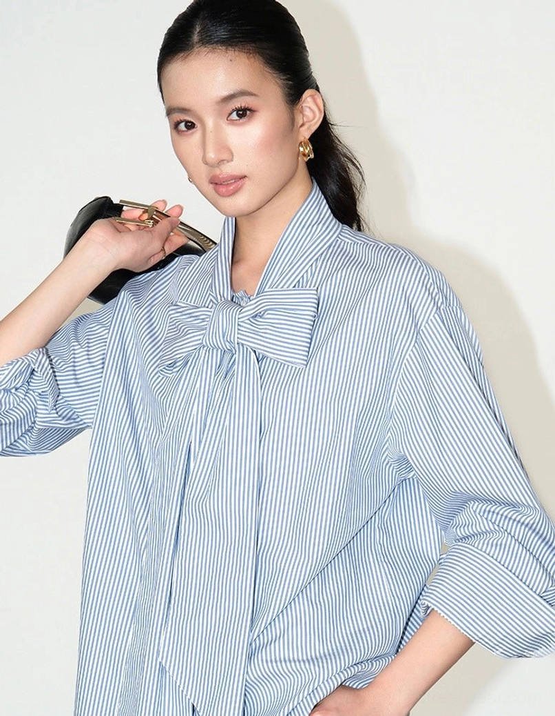 Side three-quarter view of an oversized blue pinstripe cotton blouse with a structured neck bow, relaxed sleeves, and soft Korean fashion silhouette