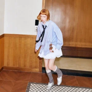 Three-quarter view of an oversized blue pinstripe shirt styled with a white layered ruffle mini skirt, black striped tie, grey knee-high socks, and white sneakers