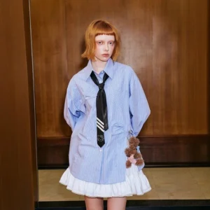 Front view of a woman wearing an oversized blue pinstripe shirt dress layered with a white ruffle mini skirt, striped tie, and teddy bear pendant in a retro elevator