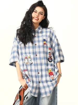 Three-quarter view of an oversized blue plaid cartoon shirt with brown buttons, curved hem, leather tote bag, and relaxed Y2K styling