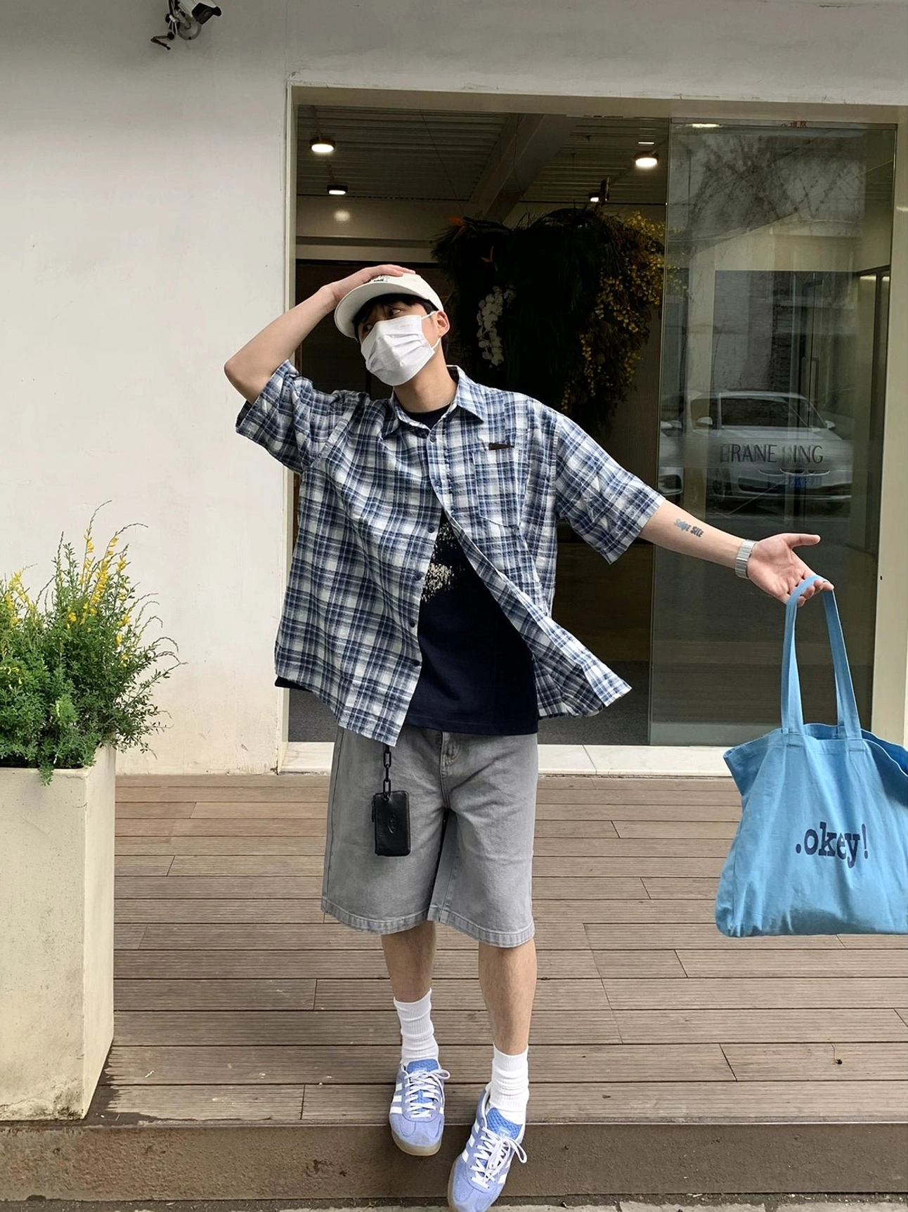 Full-body front styling of an oversized blue and white plaid short sleeve shirt with grey shorts, blue tote bag, beige cap, and light blue sneakers in a modern casual outfit