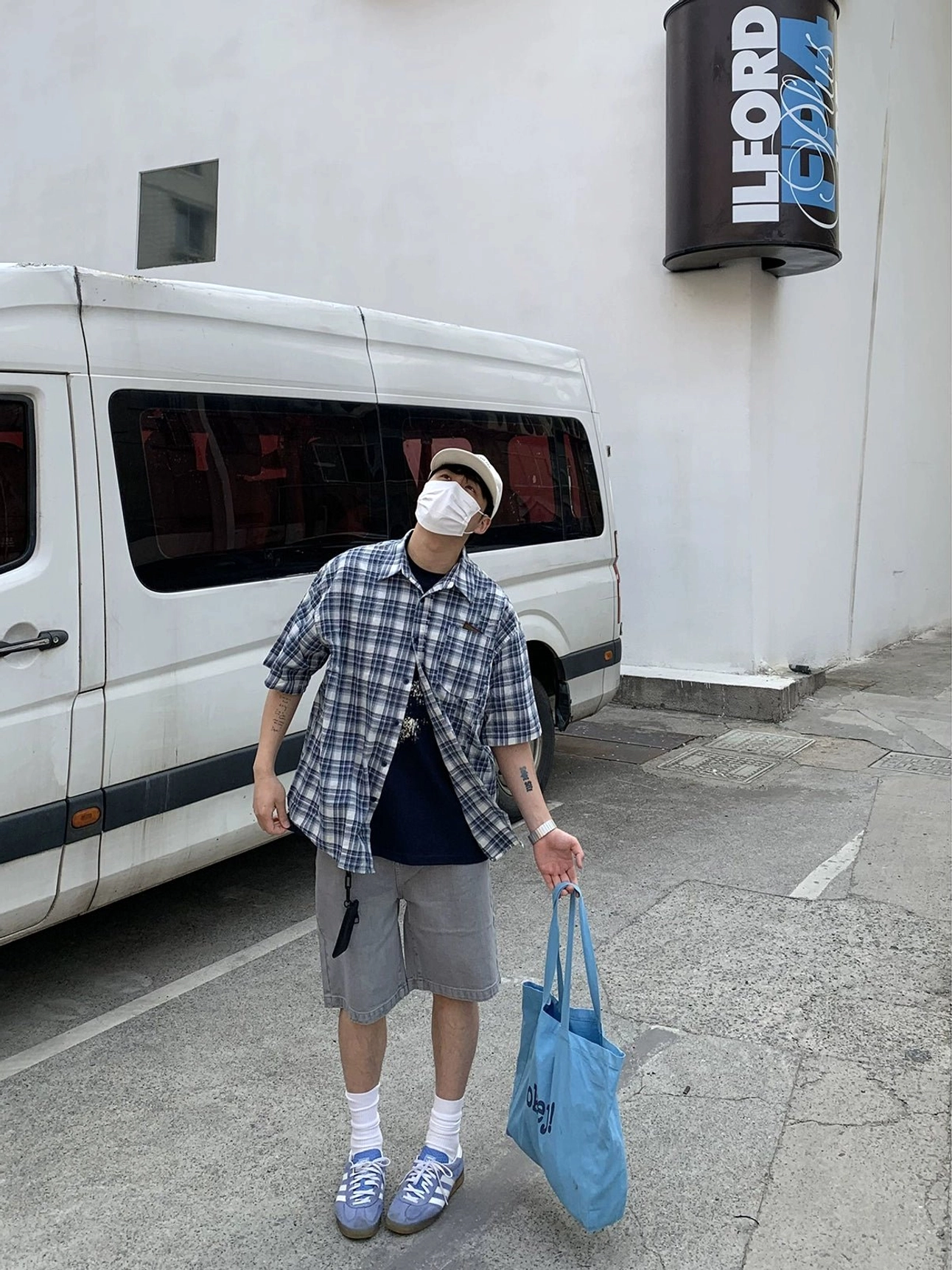 Side and back view of an oversized blue and white plaid short sleeve shirt paired with grey shorts, light blue sneakers, beige cap, and blue tote bag for Korean casual streetwear