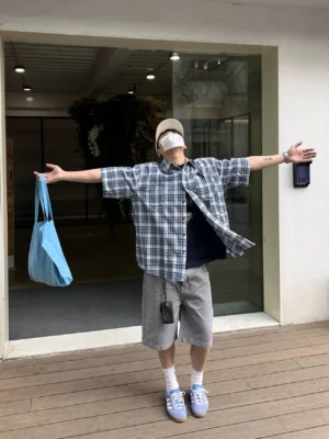 Full-body front view of an oversized blue and white plaid short sleeve shirt styled with a black T-shirt, grey shorts, light blue sneakers, beige cap, mask, and blue tote bag in a casual streetwear outfit