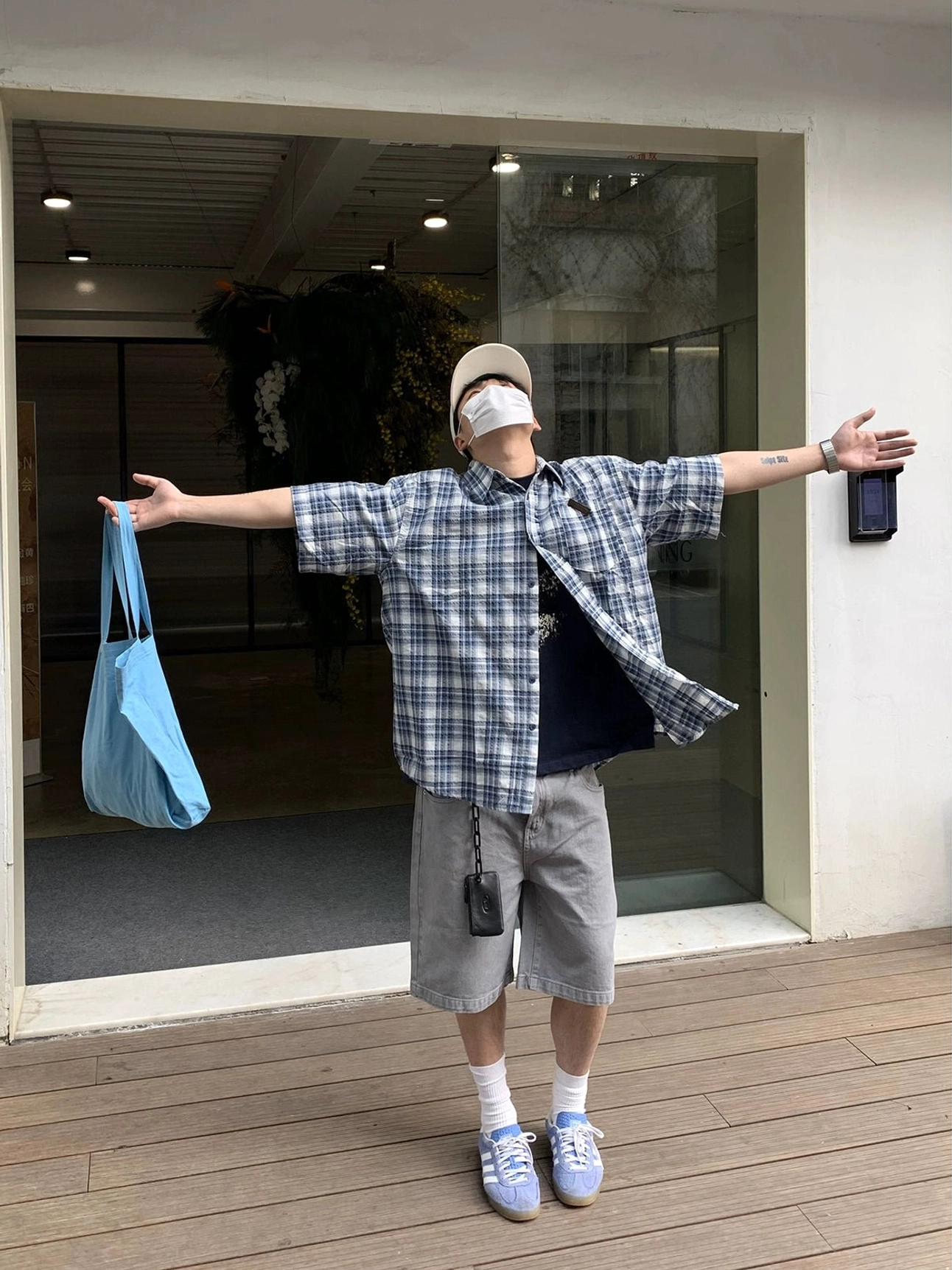 Full-body front view of an oversized blue and white plaid short sleeve shirt styled with a black T-shirt, grey shorts, light blue sneakers, beige cap, mask, and blue tote bag in a casual streetwear outfit