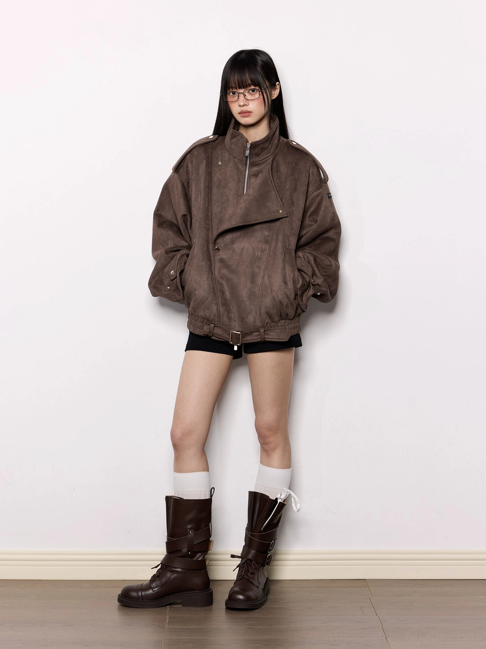Model wearing an oversized brown faux suede moto jacket with black shorts white knee-high socks and brown boots in a studio