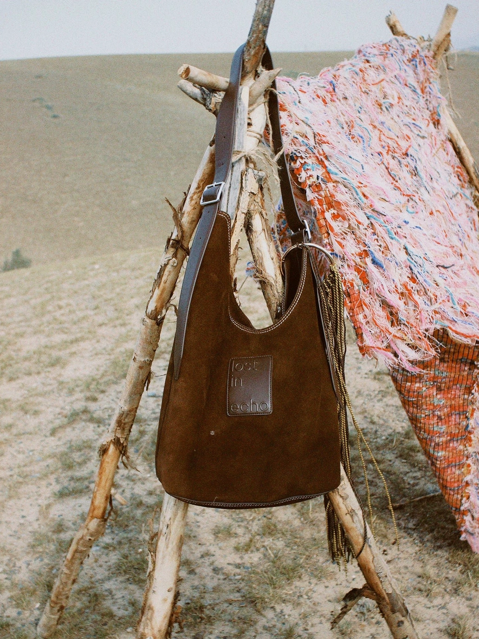 Brown suede and canvas oversized commuter tote bag hanging outdoors with adjustable shoulder strap and side tassel details
