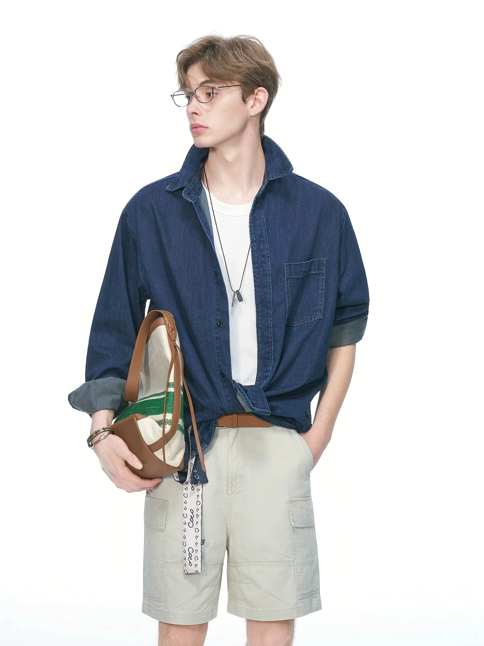 Side view of an oversized dark blue denim shirt with open collar, rolled three-quarter sleeves, chest pocket, and canvas crossbody bag