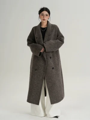 Female model wearing a black coffee and dark gray melange oversized double-breasted wool coat with wide lapels, dropped shoulders, and white wide-leg trousers in a minimalist gray studio