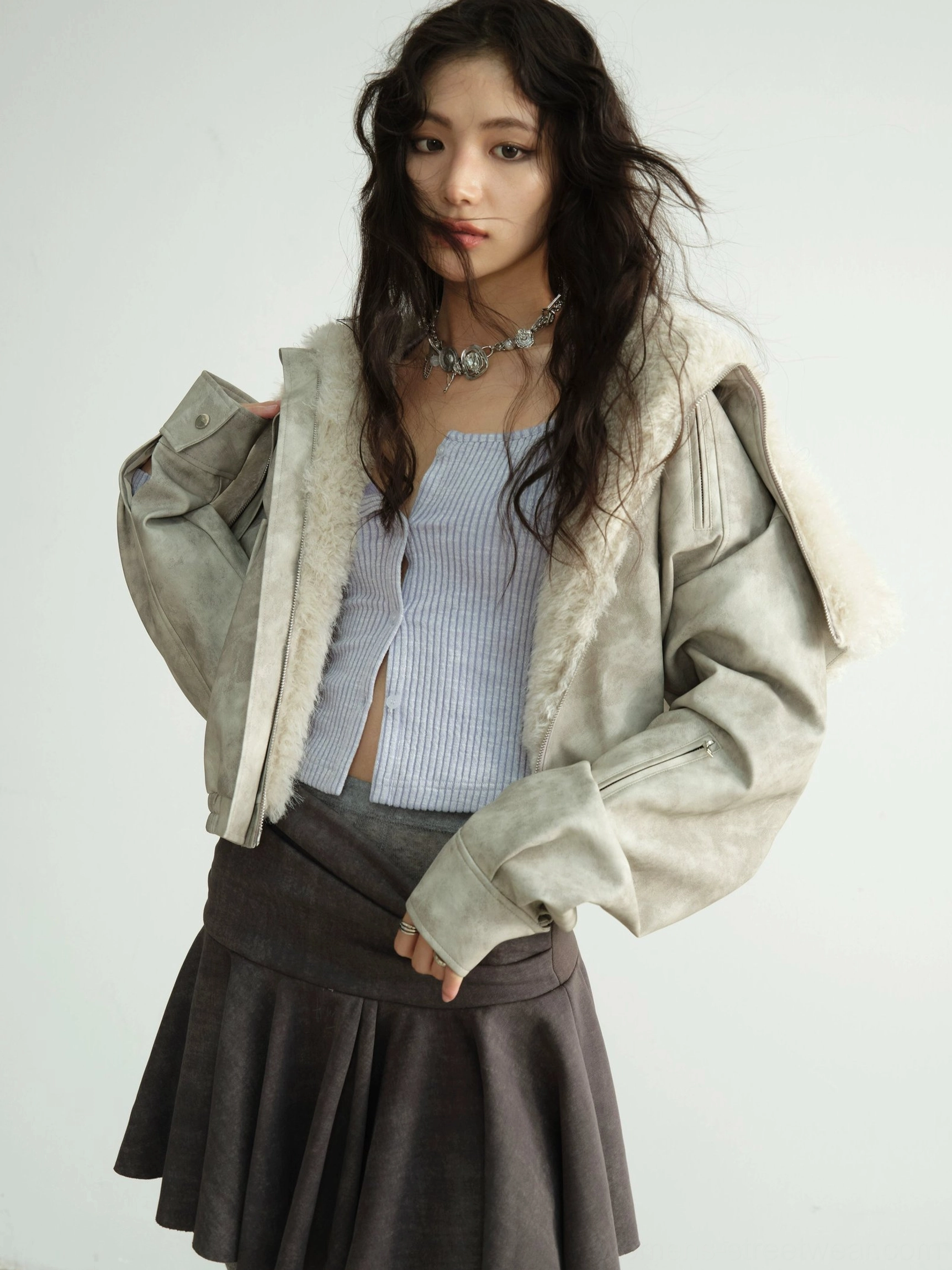 Three-quarter close-up of an oversized faux shearling hooded jacket worn open over a cropped striped top and pleated mini skirt