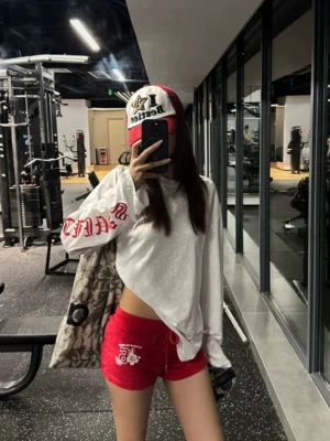 Woman wearing an oversized white gothic cross graphic long-sleeve tee with red sleeve lettering, red athletic shorts, and a baseball cap in a gym mirror selfie