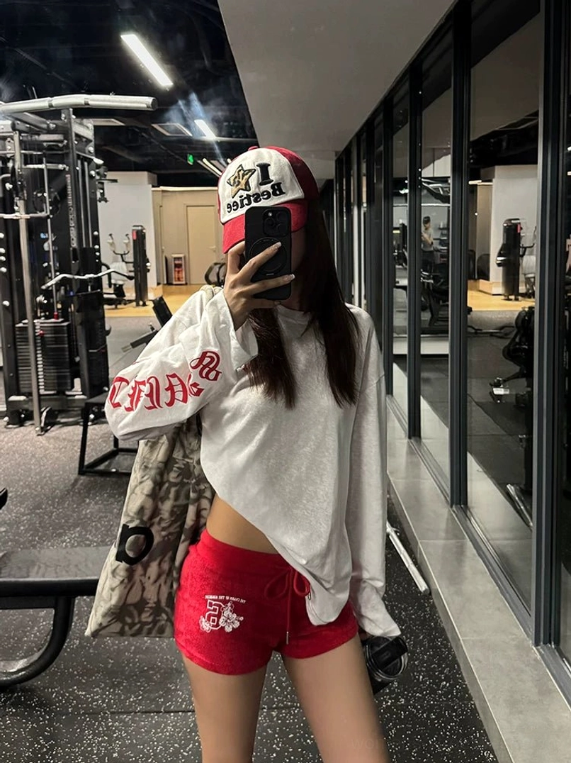 Woman wearing an oversized white gothic cross graphic long-sleeve tee with red sleeve lettering, red athletic shorts, and a baseball cap in a gym mirror selfie