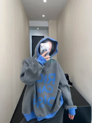 Back three-quarter view of an oversized gray streetwear hoodie showing blue reversed lettering, dropped shoulders, and contrast ribbed hem