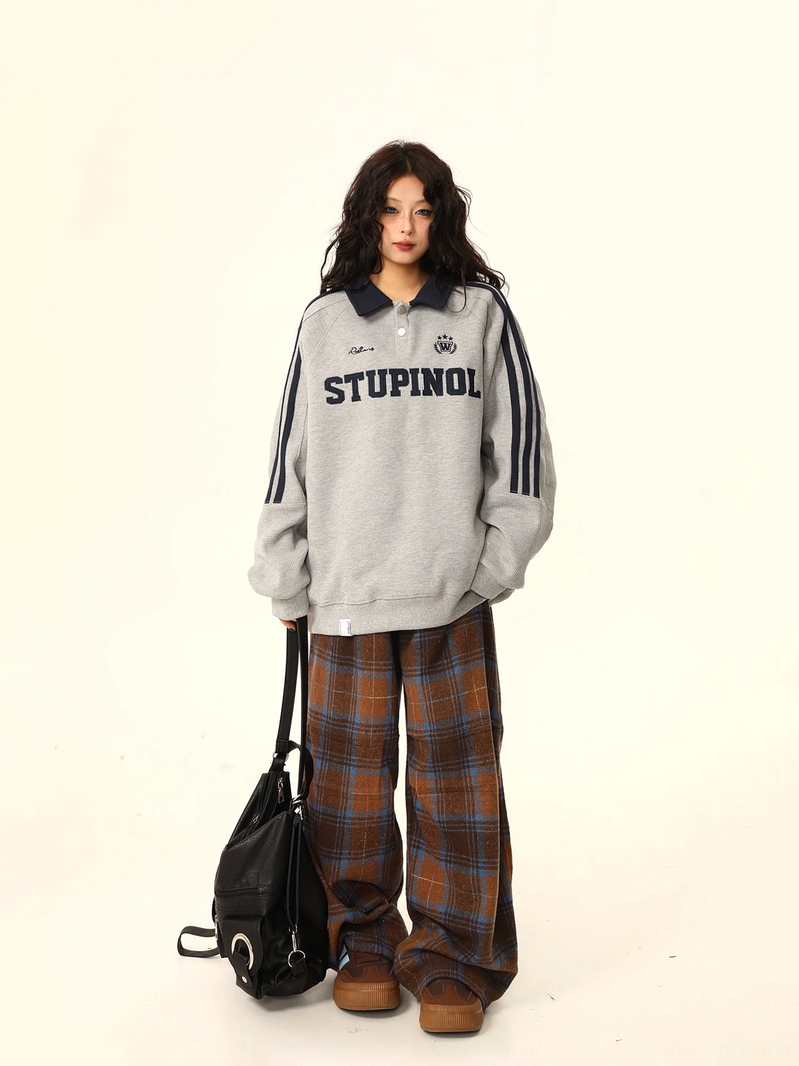 Side view of a relaxed oversized gray varsity polo sweatshirt with contrast navy details and collegiate embroidery styled with plaid pants