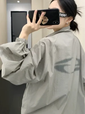 Back mirror view of an oversized gray windbreaker jacket with subtle back graphic, loose streetwear silhouette, elastic hem, long sleeves, and lightweight sun-protection design