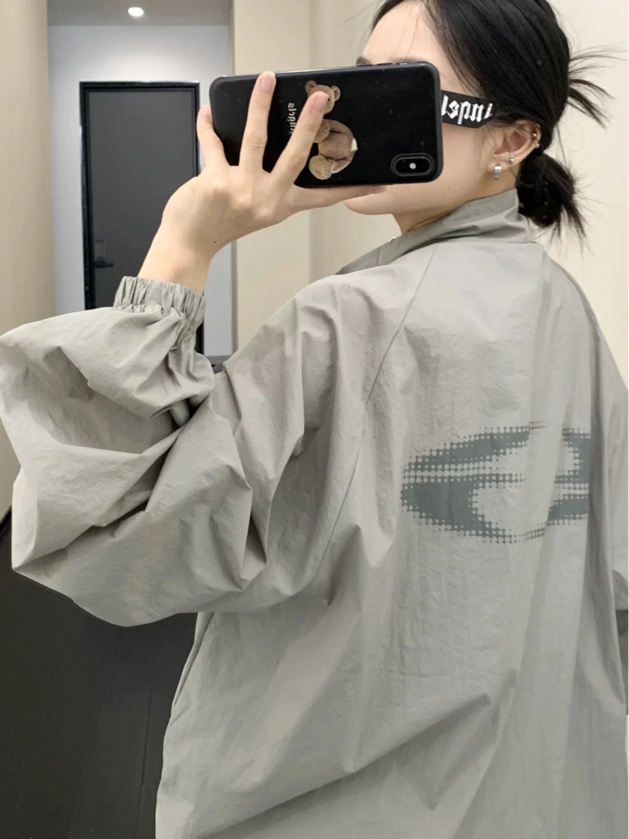 Back mirror view of an oversized gray windbreaker jacket with subtle back graphic, loose streetwear silhouette, elastic hem, long sleeves, and lightweight sun-protection design