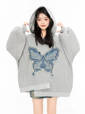 Young woman wearing an oversized grey butterfly hoodie with blue frayed butterfly chest detail and extra-long sleeves in a studio setting
