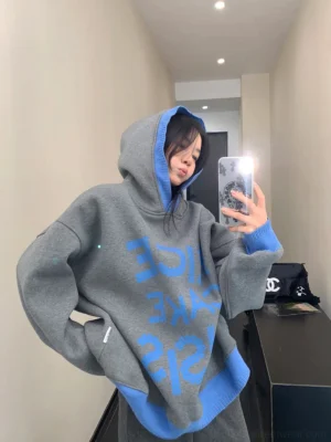 Three-quarter front view of an oversized heather gray graphic hoodie with blue ribbed trims and reversed bubble lettering in a minimalist indoor corridor