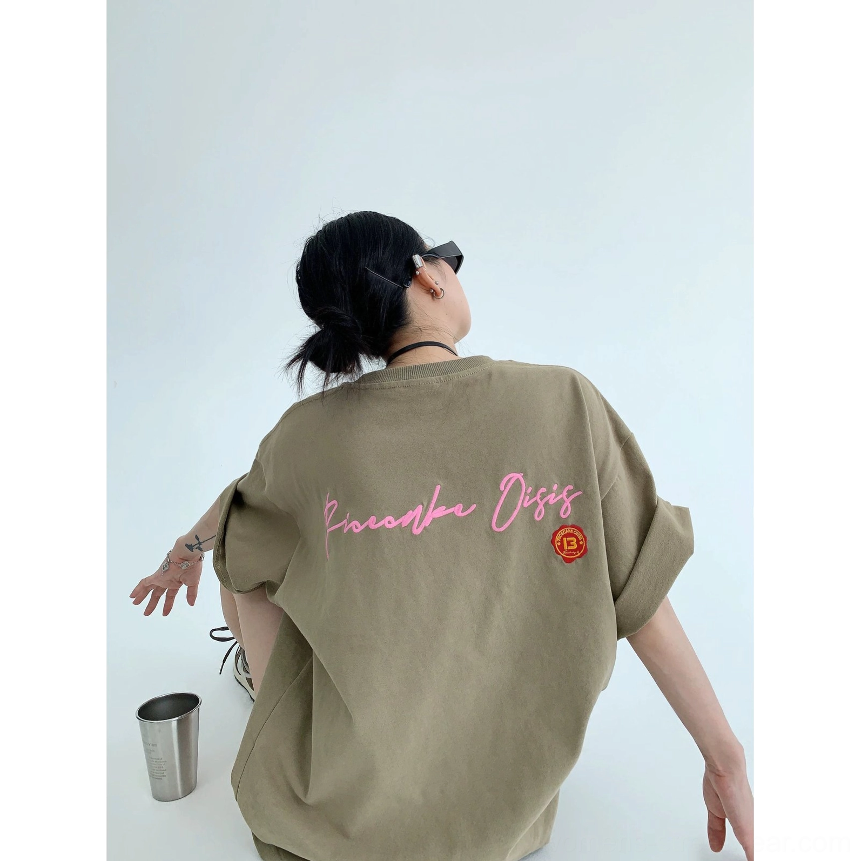 Oversized light brown cotton graphic tee shown from a low rear three-quarter angle with pink script back print and orange emblem