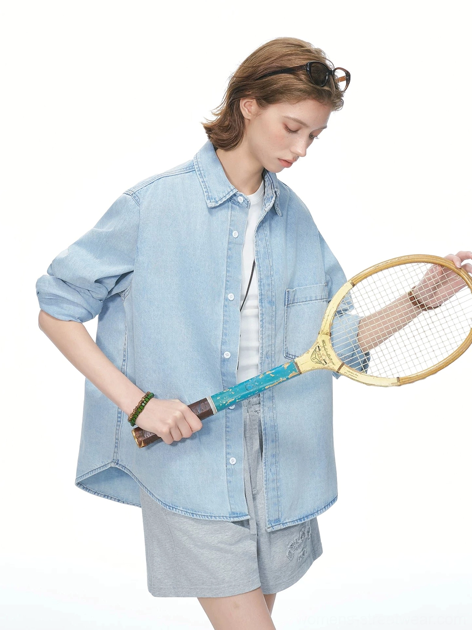 Side view of a female model in a loose oversized light wash denim shirt with half rolled sleeves and a vintage tennis racket