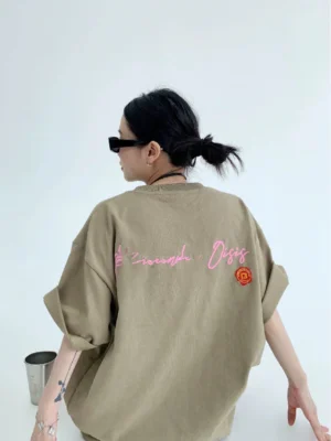 Oversized light brown neon script back graphic tee shown from a rear three-quarter view with pink cursive lettering, orange circular emblem, and rolled sleeves