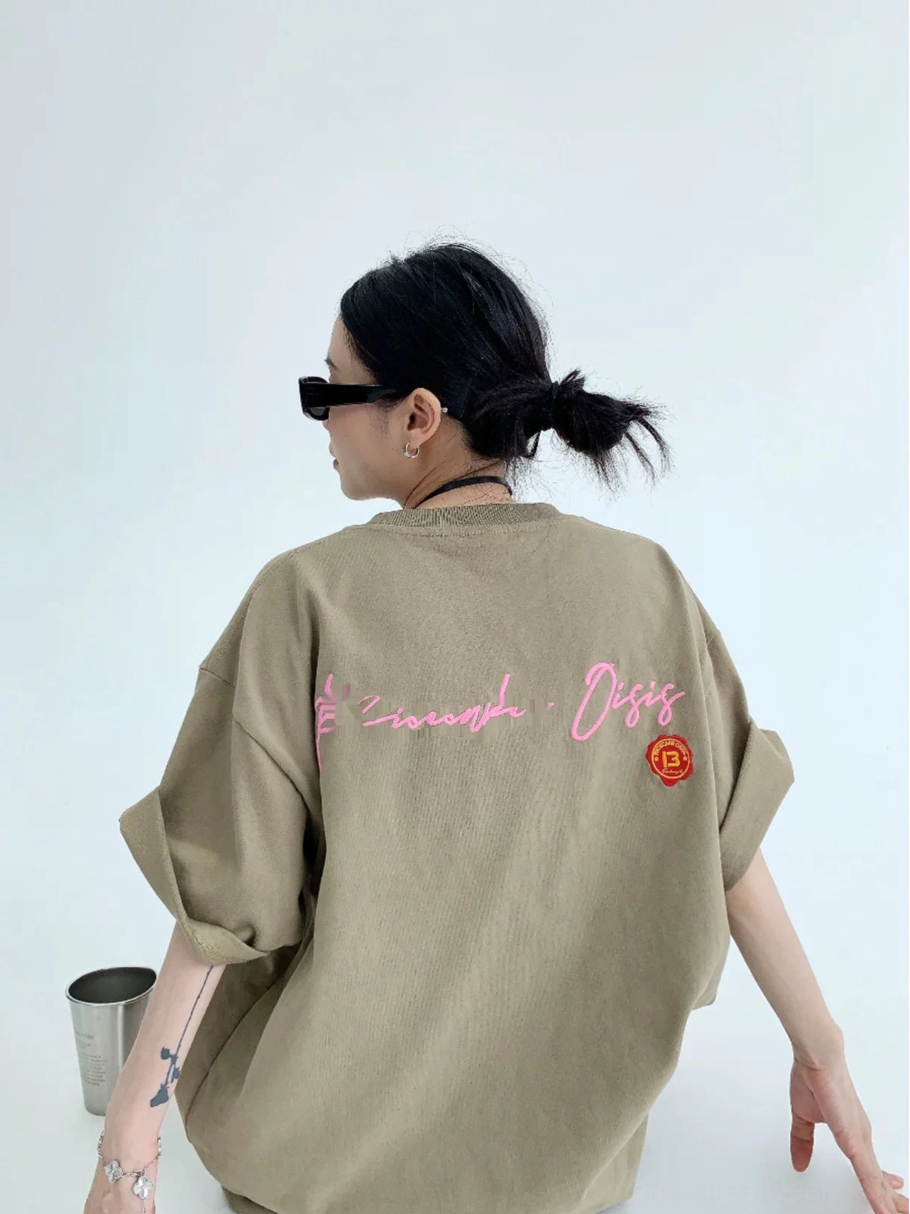 Oversized light brown neon script back graphic tee shown from a rear three-quarter view with pink cursive lettering, orange circular emblem, and rolled sleeves