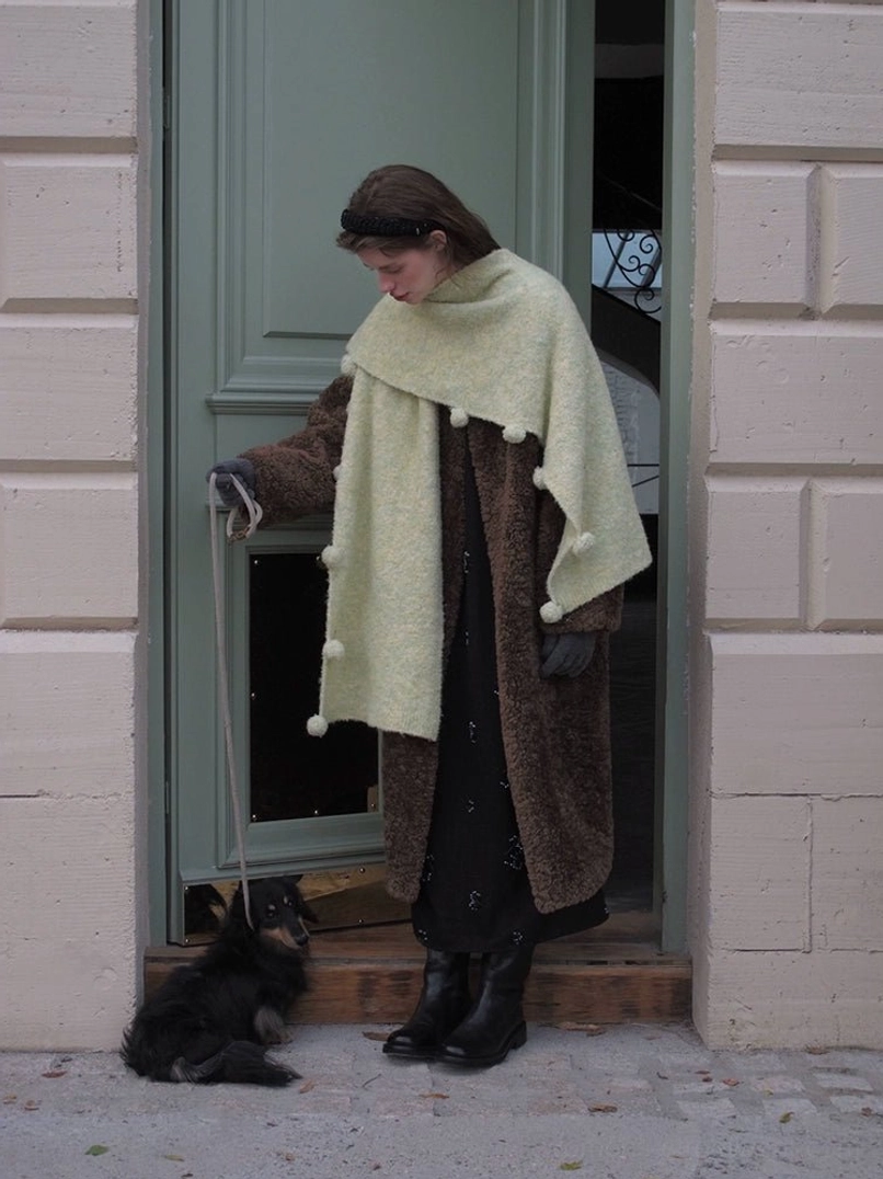 Brown women’s oversized faux shearling open-front coat styled with a light green pom-pom scarf at a doorway