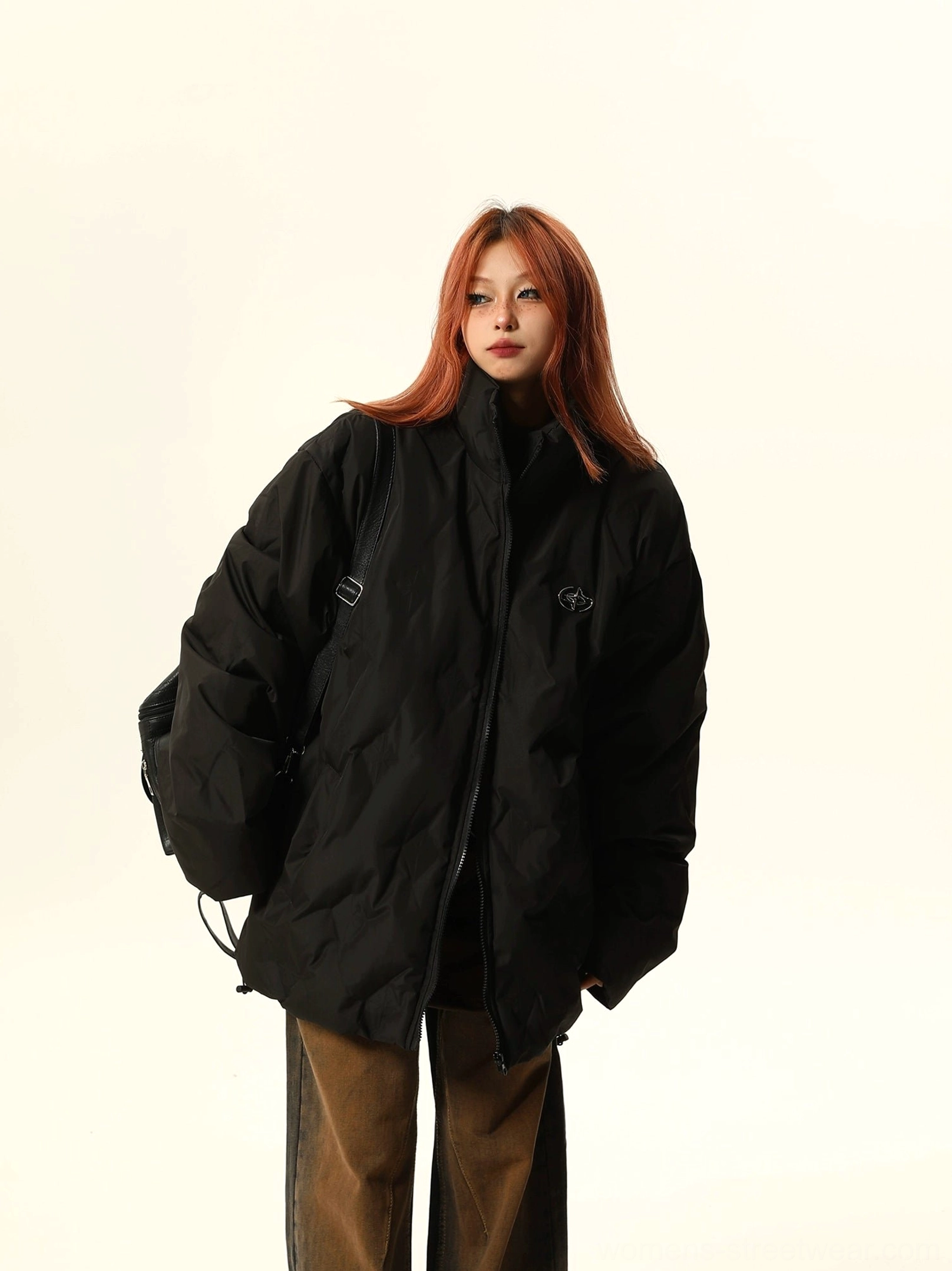 Black oversized quilted bomber jacket with stand collar, chest embroidery, hidden pockets, and adjustable drawstring hem styled with brown wide-leg pants