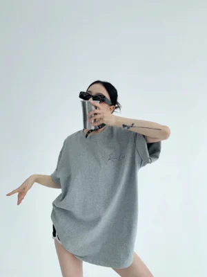 Heather gray oversized Korean streetwear cotton T-shirt front view with small cursive chest script, relaxed shoulders, sunglasses, and metal tumbler