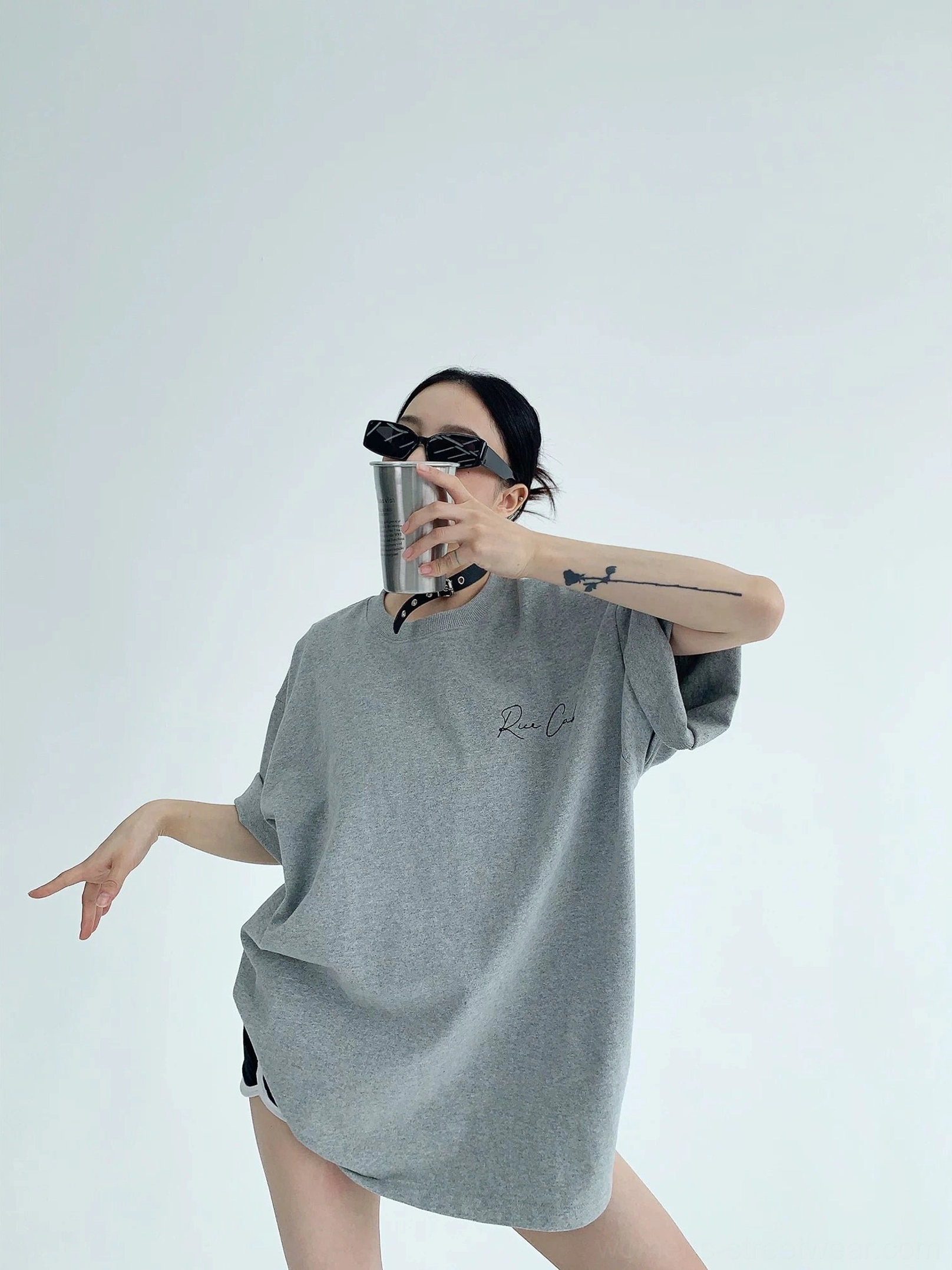 Heather gray oversized Korean streetwear cotton T-shirt front view with small cursive chest script, relaxed shoulders, sunglasses, and metal tumbler