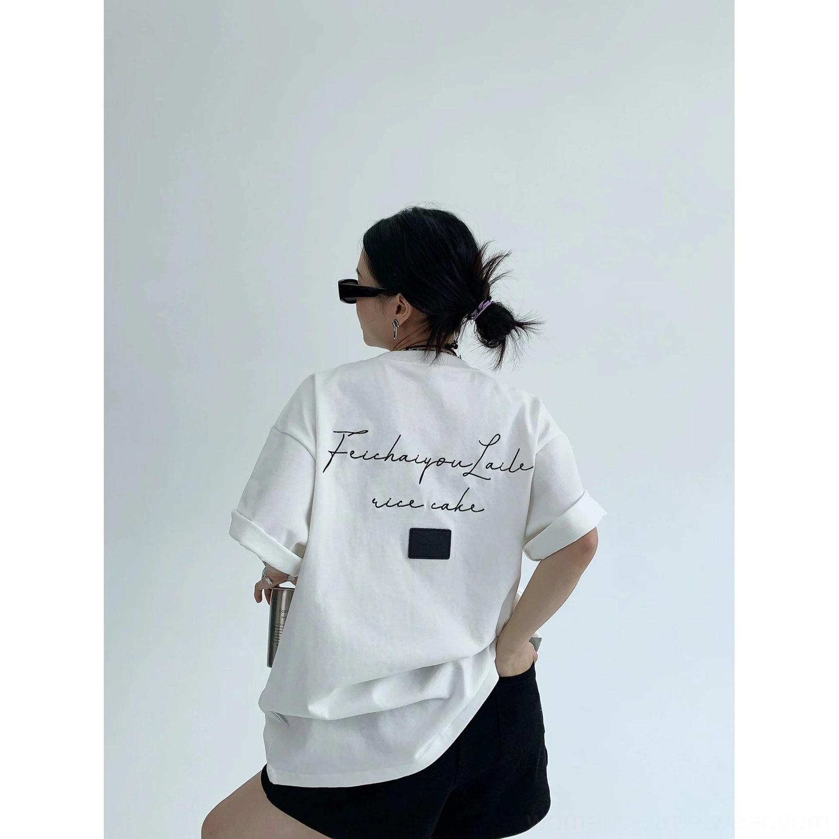 Side rear view of bright white oversized Korean streetwear T-shirt with script back design, square patch, rolled cuffs, sunglasses, and black shorts