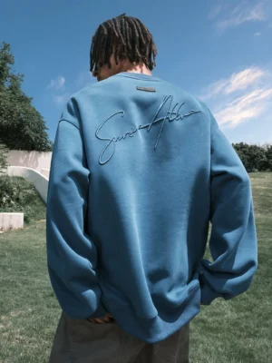 Rear view of a model wearing a dusty blue oversized script embroidered crewneck sweatshirt with dropped shoulders and raised cursive back embroidery outdoors