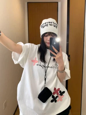 Front mirror selfie of a woman wearing a white oversized script floral graphic T-shirt with pink and black flower details, rolled sleeves, and a relaxed Korean streetwear fit
