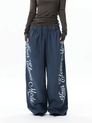 Front view of oversized blue balloon sweatpants with white vertical cursive script print on both legs, elastic drawstring waist, and wide Korean streetwear silhouette