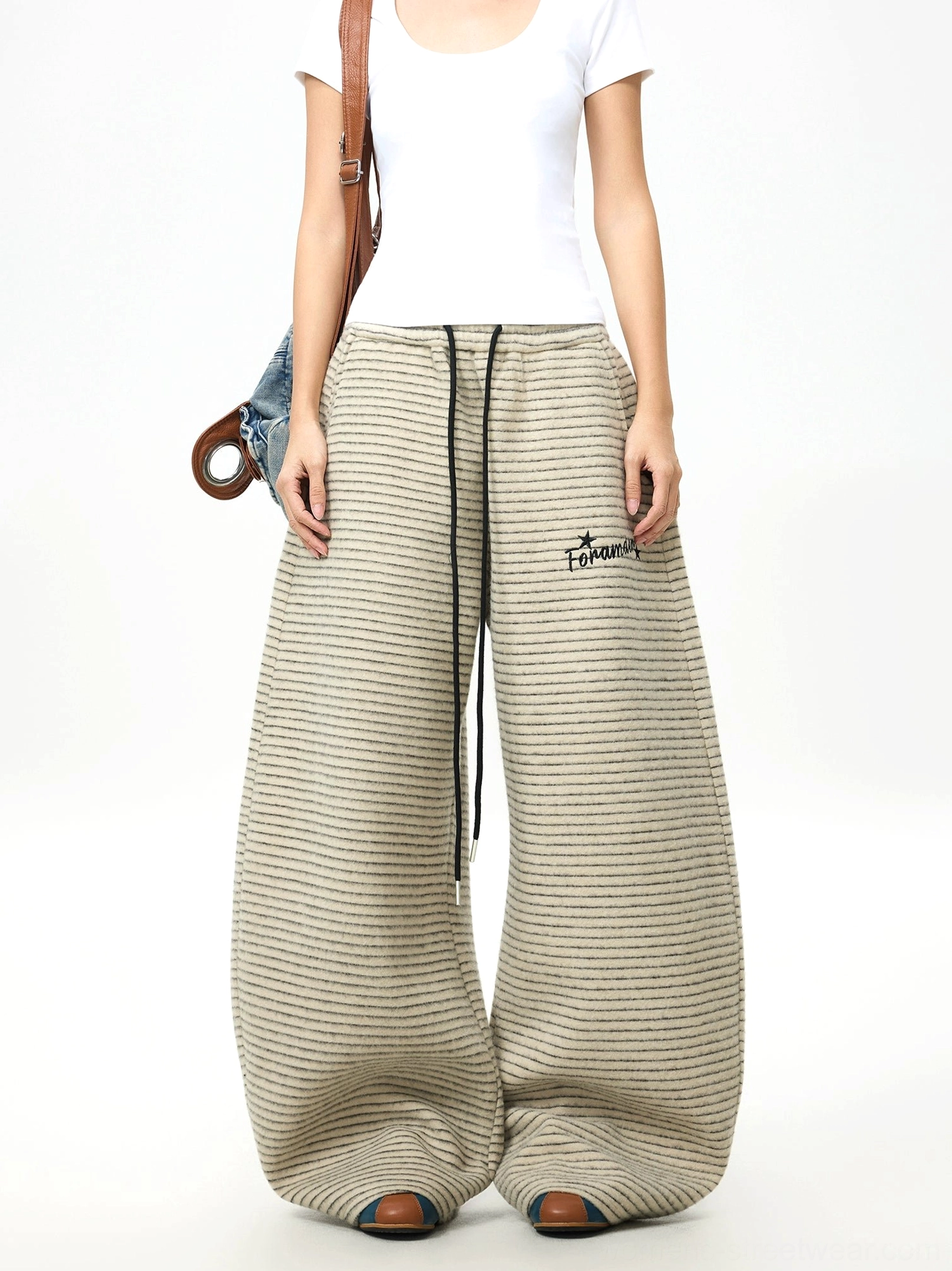 Female model styling oversized striped fleece streetwear pants with white t-shirt, flat shoes, denim accents, and crossbody bag