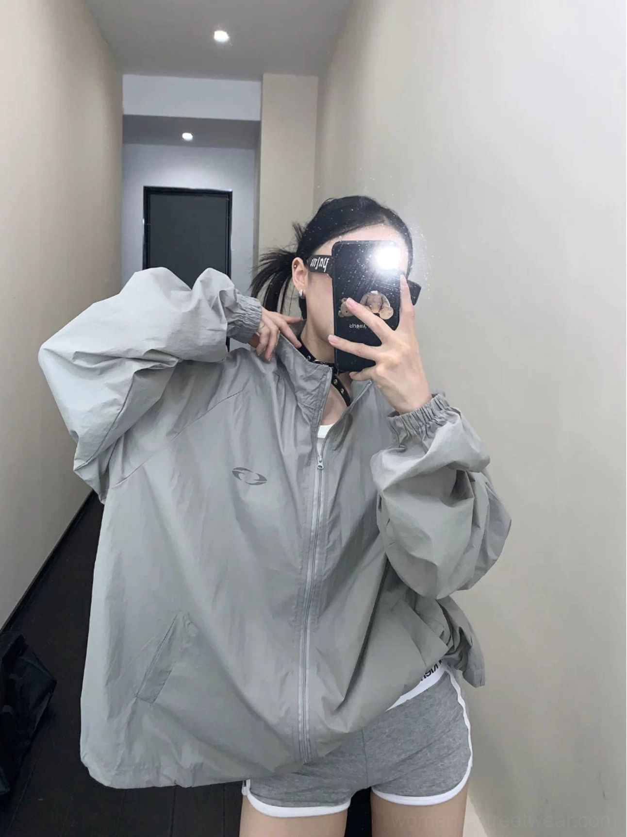 Front mirror view of a woman wearing an oversized gray UPF50+ windbreaker jacket with stand collar, full zip closure, small chest logo, elastic cuffs, and relaxed Korean streetwear fit