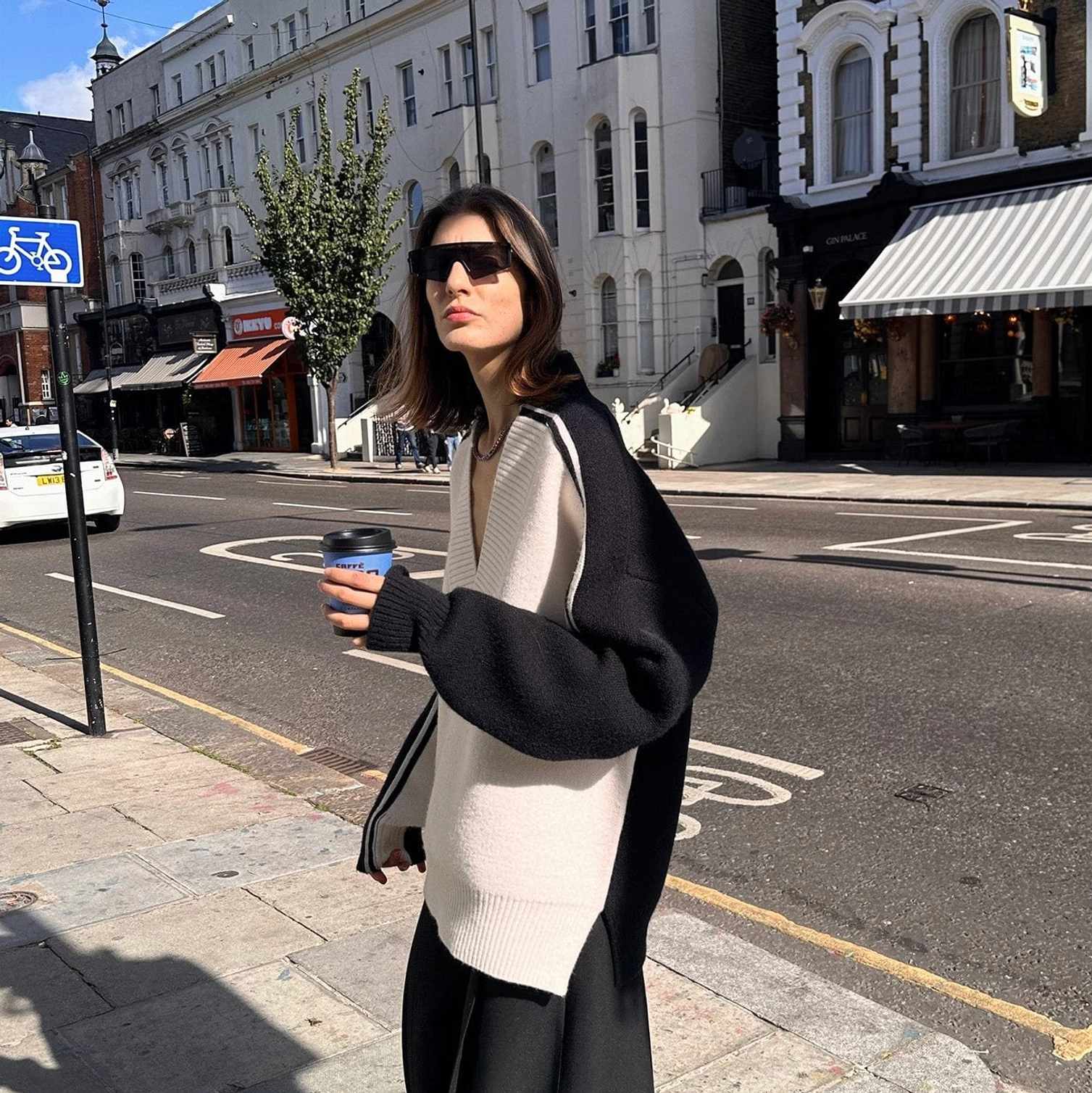 Three quarter view of relaxed oversized V-neck racing stripe sweater styled with a takeaway coffee on an urban sidewalk