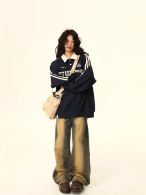 Navy blue oversized varsity polo sweatshirt with white contrast collar, athletic sleeve stripes, and embroidered collegiate lettering styled with faded wide-leg jeans