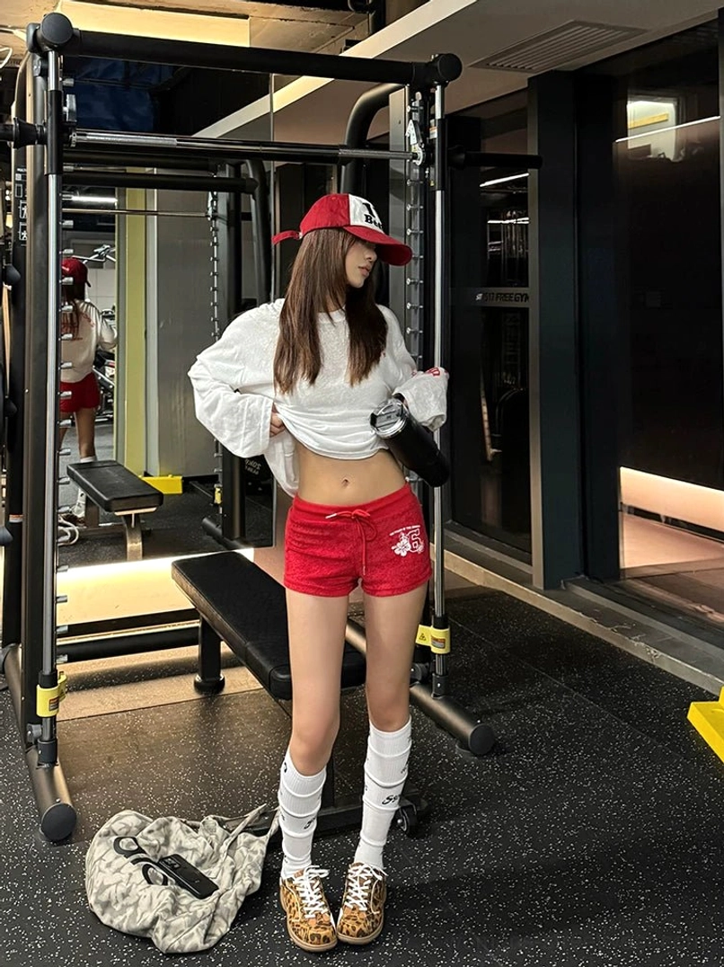 Full-body side view of an oversized white gothic cross long-sleeve streetwear tee styled with red shorts, knee-high socks, and leopard sneakers